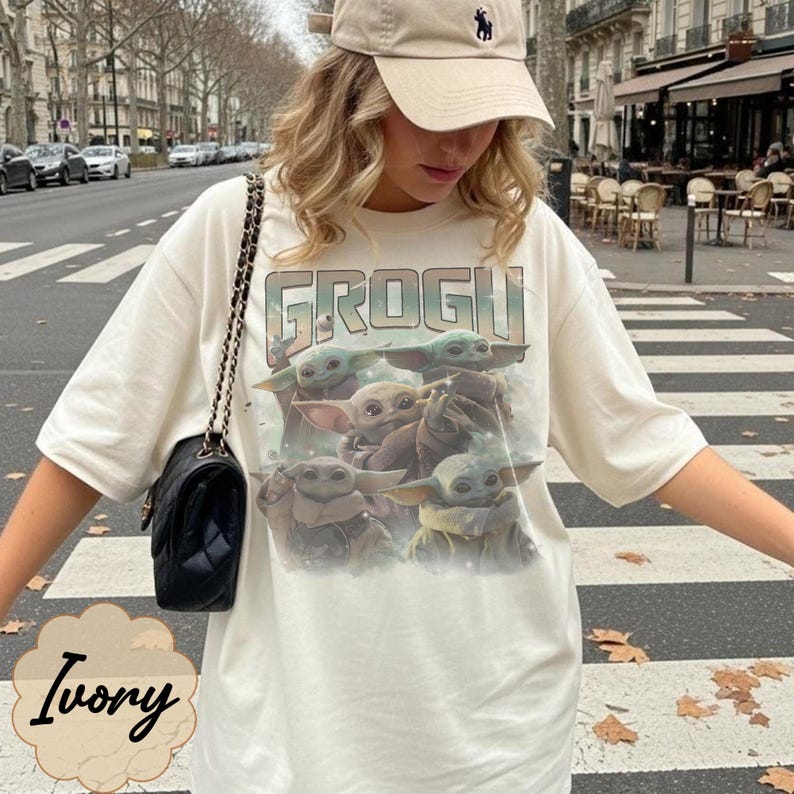 May include: An ivory short-sleeved t-shirt featuring a graphic print of multiple Grogu characters and the text GROGU in a vintage-style font. The shirt is paired with a black shoulder bag with a gold chain strap and a beige baseball cap.