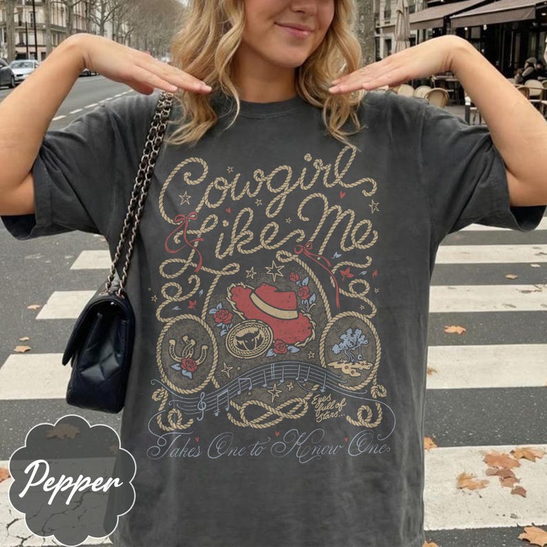 May include: A charcoal gray short-sleeve t-shirt featuring a graphic print with the text Cowgirl Like Me, a red cowboy hat, a horseshoe, a desert landscape, musical notes, and the text Eyes full of Stars and Takes One to Know One.