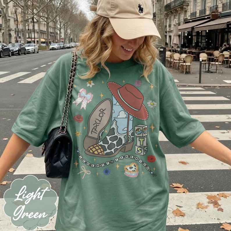 May include: A light green t-shirt featuring a graphic print of a cowboy boot with a cow print pattern, a red hat, the name Taylor, the number 13, and stacked blocks with the letters T, S, and S. The design includes small stars, flowers, and a bow.