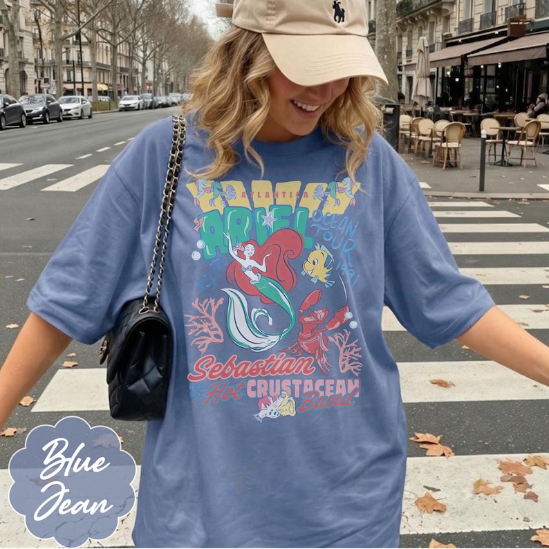 May include: Blue jean colored t-shirt featuring a graphic of Ariel, Sebastian, and Flounder from The Little Mermaid. The shirt has the words "Atlantica" and "Ocean Tour" printed on it. The shirt is a relaxed fit.