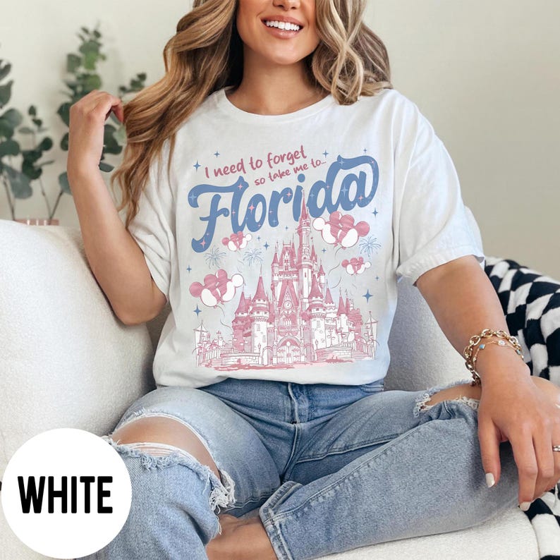 May include: White t-shirt with the text "I need to forget so take me to Florida" in blue script. The shirt features a pink and blue illustration of a castle and fireworks. The shirt is a casual style.