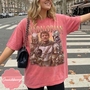 Pu&ograve; includere: Una t-shirt a maniche corte color corallo con una stampa grafica dei personaggi di The Mandalorian. Il design include il testo "MANDALORIAN" in un carattere dorato sopra una figura centrale circondata da personaggi corazzati e una creatura.