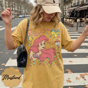 May include: Mustard-coloured t-shirt with a graphic of Ariel from Disney's The Little Mermaid. The design includes Ariel with pink hair, Flounder, and other characters. The shirt has short sleeves and a crew neck.