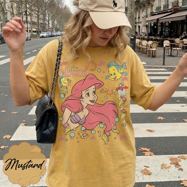 May include: Mustard-coloured t-shirt with a graphic of Ariel from Disney's The Little Mermaid. The design includes Ariel with pink hair, Flounder, and other characters. The shirt has short sleeves and a crew neck.