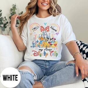 May include: White t-shirt with a colorful graphic featuring Disney characters, a castle, and the text "My First Disney Trip." The design includes a rainbow, a bow, and the word "Name."