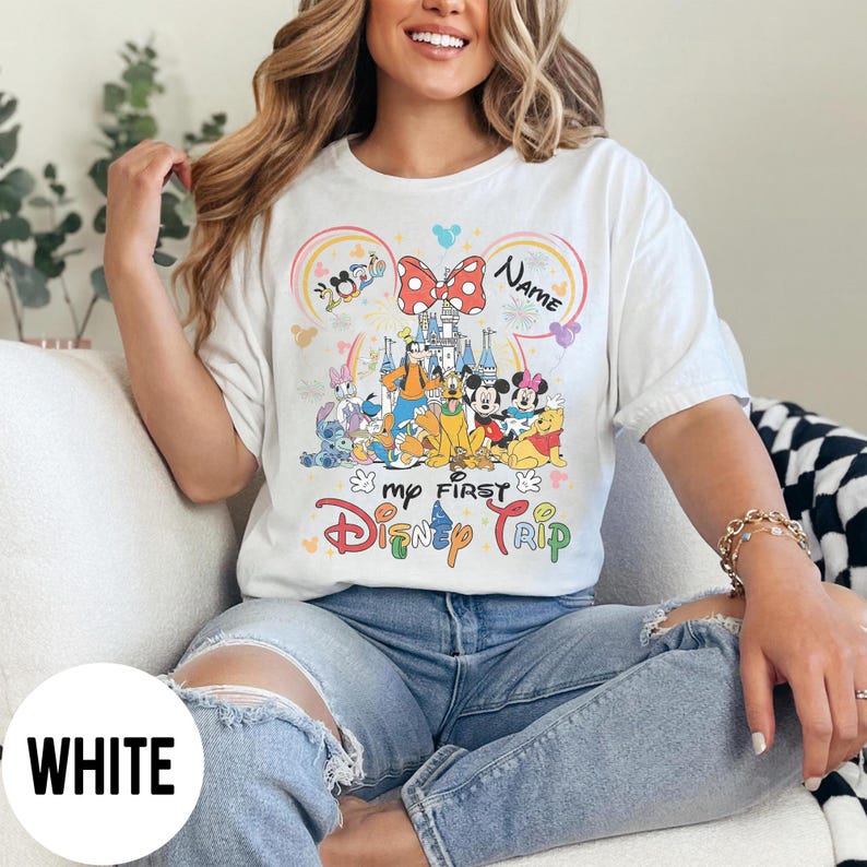 May include: White t-shirt with a colorful graphic featuring Disney characters, a castle, and the text "My First Disney Trip." The design includes a rainbow, a bow, and the word "Name."