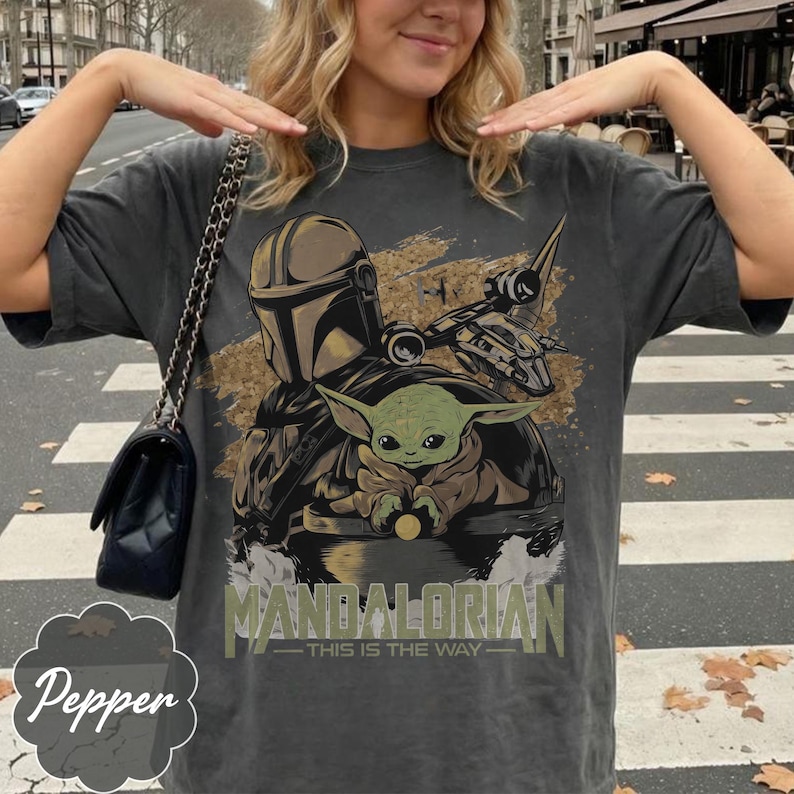Może przedstawiać: Ciemnoszara koszulka z grafiką przedstawiającą The Mandalorian, Grogu i statek kosmiczny. Tekst "MANDALORIAN - THIS IS THE WAY" jest wydrukowany poniżej. Koszulka ma wygląd vintage.