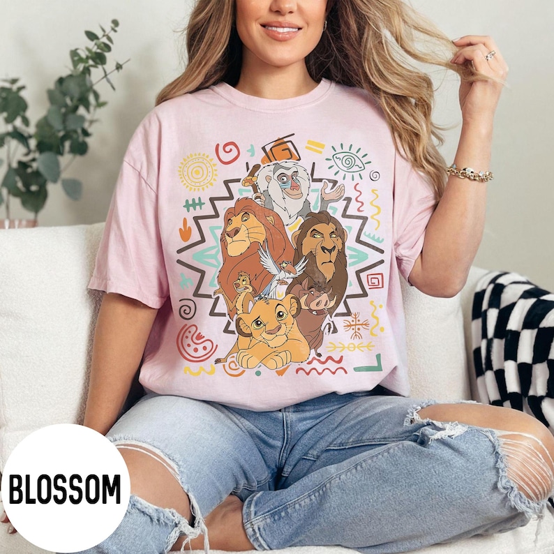 Puede incluir: Camiseta rosa claro con un dise&ntilde;o gr&aacute;fico colorido de personajes de El Rey Le&oacute;n. El dise&ntilde;o incluye a Simba, Mufasa, Scar, Tim&oacute;n, Pumba y Rafiki, rodeados de patrones tribales. La palabra "BLOSSOM" est&aacute; impresa en la parte inferior izquierda.