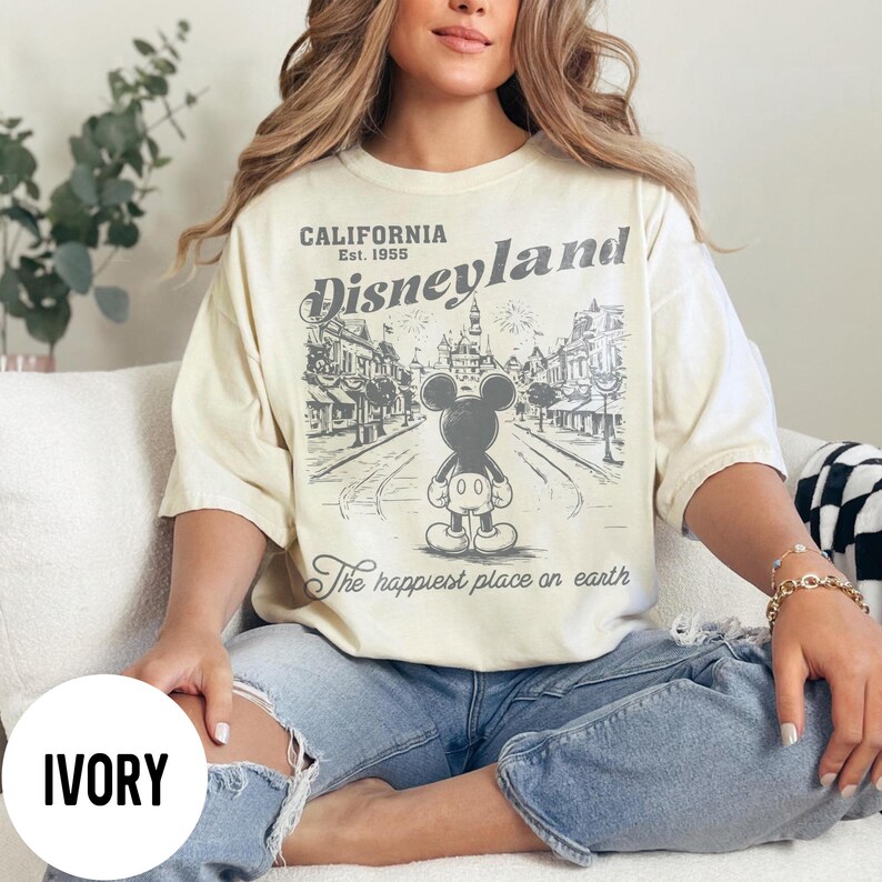 Może przedstawiać: Kremowa koszulka z grafiką Disneyland. Projekt zawiera napis "CALIFORNIA Est. 1955 Disneyland" nad ilustracją Myszki Miki. Poniżej znajduje się fraza "The happiest place on earth". Koszulka jest noszona z niebieskimi dżinsami.
