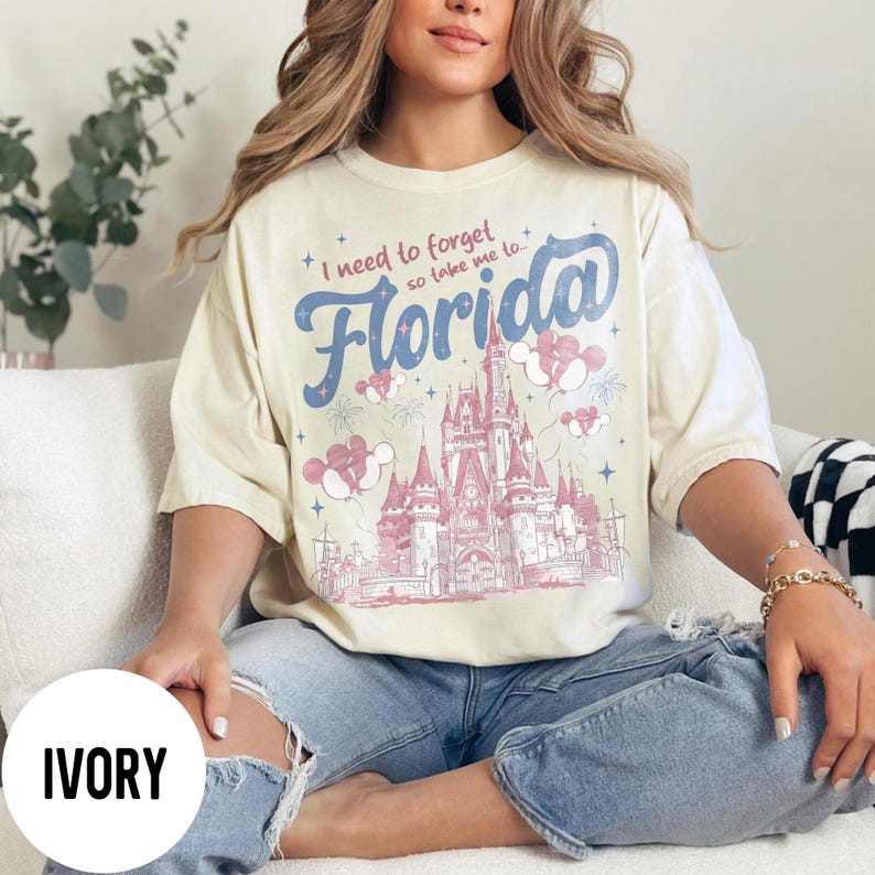 May include: Ivory-colored t-shirt featuring a graphic of a castle with the text "I need to forget so take me to Florida". The design includes pink and blue accents, with Mickey Mouse balloons and fireworks.