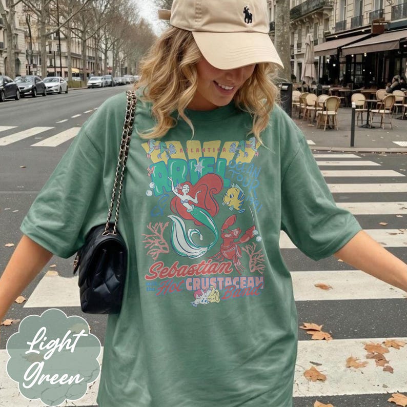 May include: Light green t-shirt featuring a graphic of Ariel, Flounder, and Sebastian from The Little Mermaid. The shirt has the words "Sebastian Crustacean" and "Light Green" printed on it. The shirt is a relaxed fit.