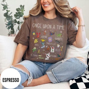 May include: Espresso-coloured t-shirt featuring the text "ONCE UPON A TIME" and illustrations of cartoon characters. The design includes Jasmine, Ariel, Cinderella, and Belle. The word "ESPRESSO" is printed on the bottom left of the shirt.