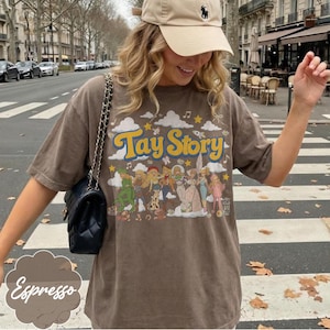May include: A brown t-shirt featuring the text Tay Story in yellow letters. The graphic includes a series of illustrated figures in various outfits, surrounded by clouds, stars, and musical notes. A black quilted shoulder bag with a chain strap is worn.