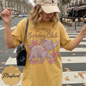 Op de afbeelding: Mosterdkleurig T-shirt met een cartoon olifantenontwerp en de tekst "Lumpy's Garden Club". De olifant is paars met roze bloemen. Het shirt heeft een relaxte pasvorm.