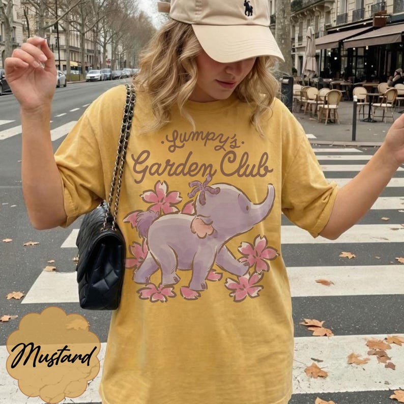 Op de afbeelding: Mosterdkleurig T-shirt met een cartoon olifantenontwerp en de tekst "Lumpy's Garden Club". De olifant is paars met roze bloemen. Het shirt heeft een relaxte pasvorm.