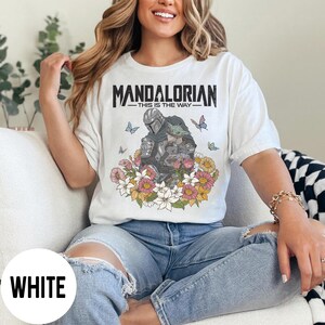 Puede incluir: Camiseta blanca con la frase "MANDALORIAN - THIS IS THE WAY" y un gr&aacute;fico del Mandaloriano y Grogu, rodeados de flores y mariposas. La camiseta tiene un corte holgado.