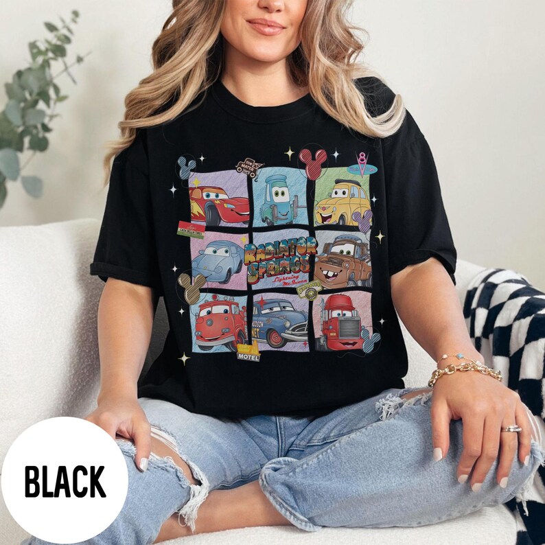 May include: Black t-shirt featuring a grid of cartoon cars from the movie Cars, with the text "Radiator Springs" in the center. The design includes various car characters in colorful squares. The word "BLACK" is printed on a white circle.