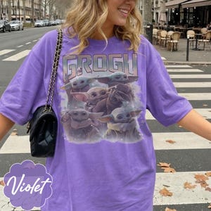 May include: A violet short-sleeved t-shirt featuring a graphic print of four Grogu characters from The Mandalorian. The text GROGU is printed in large block letters above the characters. A black quilted shoulder bag with a chain strap is visible.