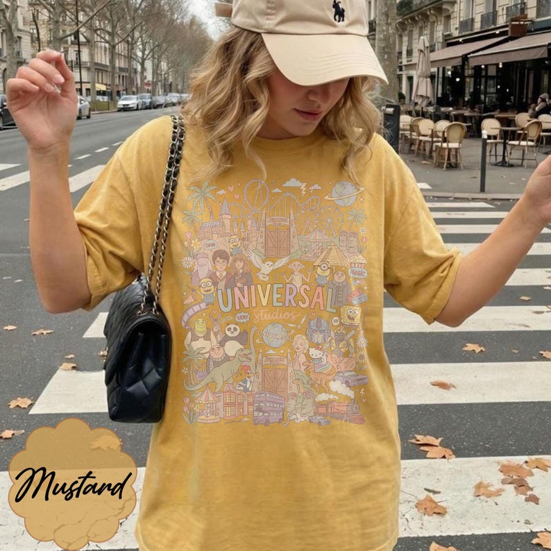 May include: Mustard yellow t-shirt featuring a colourful graphic of Universal Studios attractions and characters. The shirt has the text "UNIVERSAL Studios". The t-shirt is short-sleeved and casual.