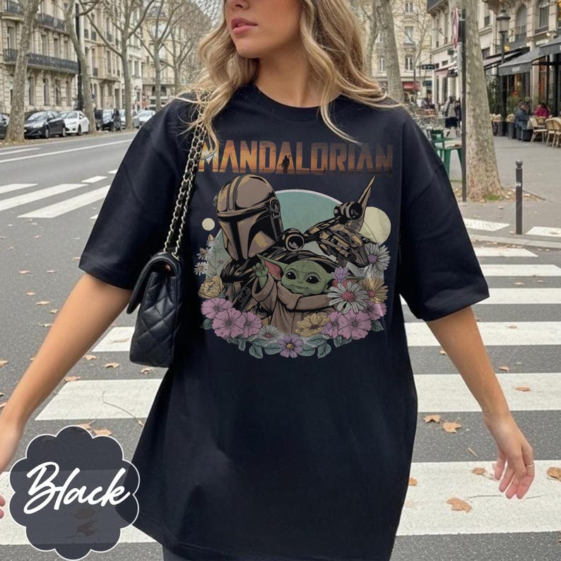 May include: Black t-shirt featuring a graphic of The Mandalorian and Grogu surrounded by flowers. The text "MANDALORIAN" is printed across the top. The shirt is a short-sleeved, oversized style.