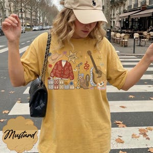 May include: A mustard yellow t-shirt featuring a graphic print of a cowboy hat, a red paisley cardigan, a sheriff badge labeled "Sheriff Taylor," a microphone, a cowboy boot, a guitar, and blocks spelling "Toy Story" with "Taylor" on the boot.