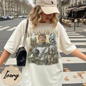 Pu&ograve; includere: Una t-shirt color avorio a maniche corte con una stampa grafica dei personaggi di The Mandalorian, tra cui una figura centrale, diversi guerrieri in armatura e una piccola creatura verde. La maglia ha un girocollo e una vestibilit&agrave; ampia.