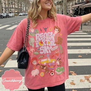 Pode incluir: Uma camiseta coral com um design gr&aacute;fico colorido com o texto "Reach for the Sky" e v&aacute;rios personagens de desenhos animados. O design inclui um cowboy, um cacto e um caminh&atilde;o de brinquedo. A camisa &eacute; um estilo casual de manga curta.