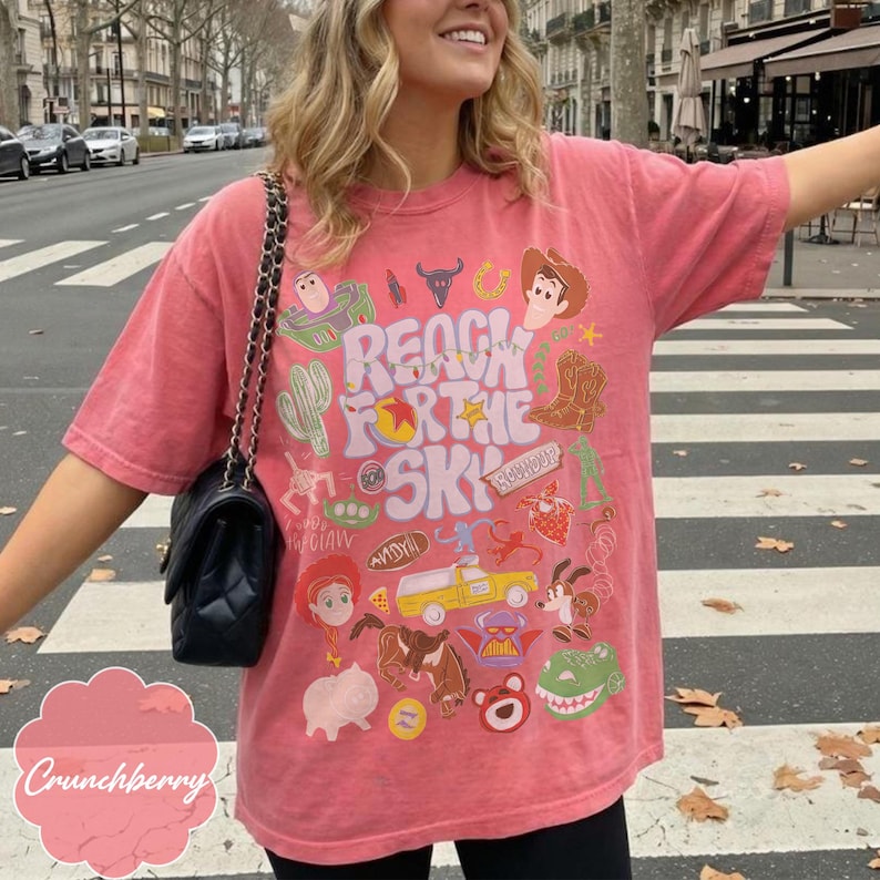 Pode incluir: Uma camiseta coral com um design gr&aacute;fico colorido com o texto "Reach for the Sky" e v&aacute;rios personagens de desenhos animados. O design inclui um cowboy, um cacto e um caminh&atilde;o de brinquedo. A camisa &eacute; um estilo casual de manga curta.