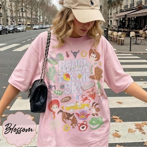 Pode incluir: Camiseta rosa claro com a frase "Reach for the Sky" e v&aacute;rios gr&aacute;ficos de personagens de desenhos animados. A camiseta tem gola redonda e mangas curtas. A palavra "Blossom" est&aacute; na parte inferior esquerda.