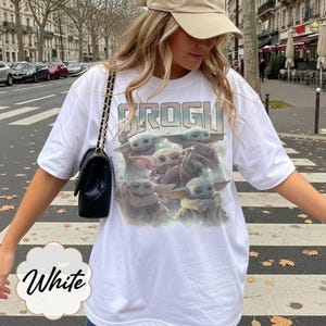 May include: A white short-sleeved t-shirt featuring a graphic print of four green creatures with large ears and eyes. The text "GROGU" is printed in a bold, distressed font above the characters. The shirt has a crew neckline and a loose fit.