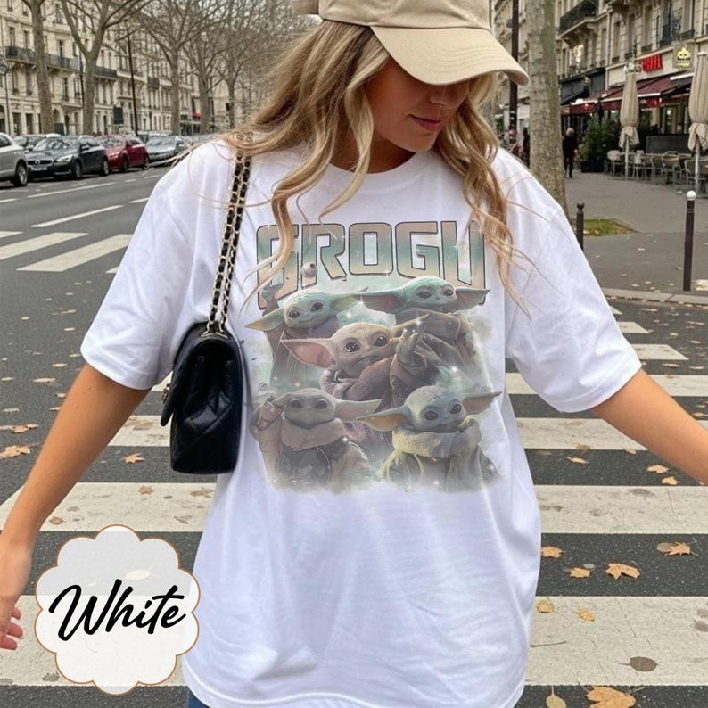 May include: A white short-sleeved t-shirt featuring a graphic print of four green creatures with large ears and eyes. The text "GROGU" is printed in a bold, distressed font above the characters. The shirt has a crew neckline and a loose fit.