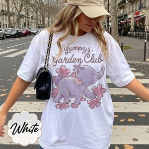 Op de afbeelding: Wit T-shirt met een paarse olifant, roze bloemen en de tekst "Gumpy's Garden Club." Het shirt heeft korte mouwen en een losse pasvorm. Het woord "White" is geschreven in een wit wolk-vormig ontwerp.