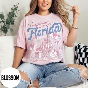 May include: A light pink t-shirt with the text "I need to forget so take me to Florida" in blue script. The shirt features a castle graphic with Mickey Mouse balloons and fireworks.