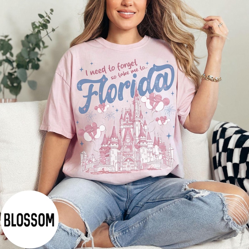 May include: A light pink t-shirt with the text "I need to forget so take me to Florida" in blue script. The shirt features a castle graphic with Mickey Mouse balloons and fireworks.