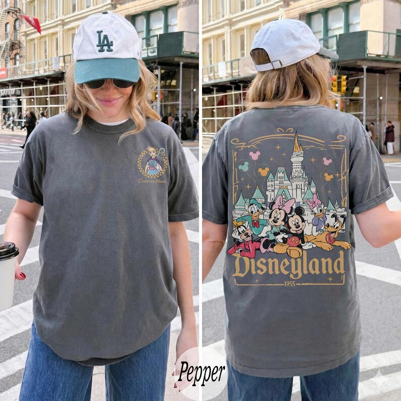 Pu&ograve; includere: T-shirt color pepe con grafica Disneyland sul retro, con personaggi dei cartoni animati e un castello. La parte anteriore ha un design pi&ugrave; piccolo. La maglietta &egrave; indossata con un berretto bianco e jeans blu.