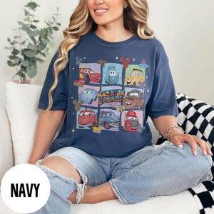 May include: Navy blue t-shirt featuring a colorful graphic of cartoon cars in a grid pattern. The graphic includes the text "Radiator Springs" and other car-related imagery. The shirt is a comfortable, casual style.