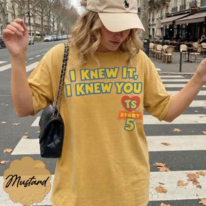 May include: A mustard yellow short-sleeve t-shirt featuring the blue text I KNEW IT, I KNEW YOU, a red heart with the letters TS, and the number 5 in blue. The shirt is paired with a black quilted shoulder bag with a gold chain strap.