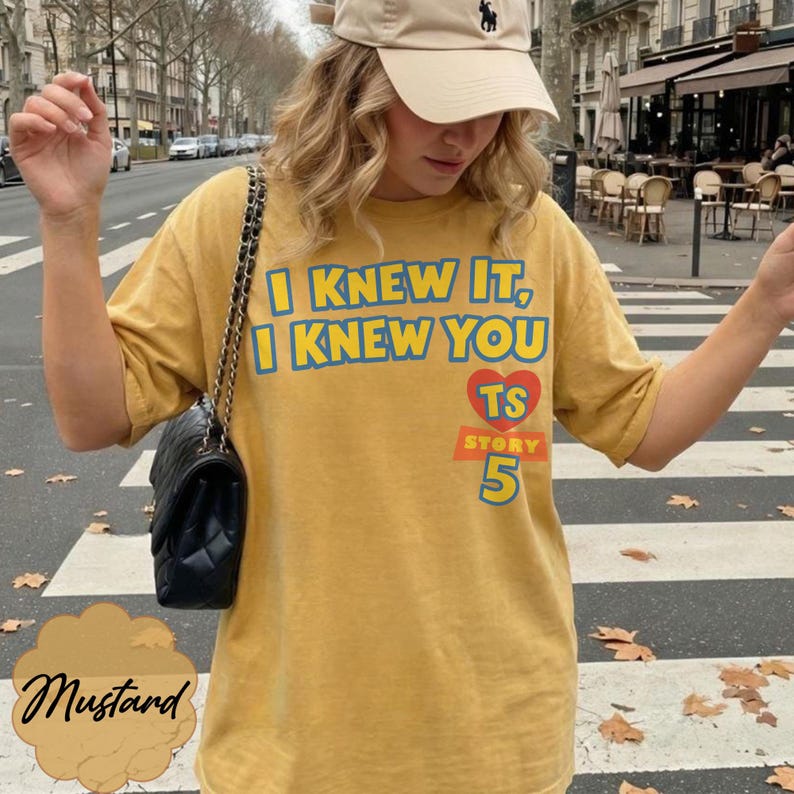 May include: A mustard yellow short-sleeve t-shirt featuring the blue text I KNEW IT, I KNEW YOU, a red heart with the letters TS, and the number 5 in blue. The shirt is paired with a black quilted shoulder bag with a gold chain strap.