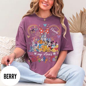 May include: Berry-colored t-shirt featuring a colorful Disney graphic. The design includes a castle, fireworks, and various Disney characters. Text on the shirt reads "my first Disney trip" and "Name".