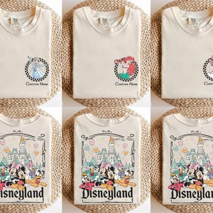 Pu&ograve; includere: T-shirt color crema con disegni di personaggi Disney. Alcune magliette presentano Elsa, Ariel e Paperina in cornici circolari a scacchi con il testo "Custom Name". Altre magliette mostrano una grafica di Disneyland con vari personaggi e la scritta "Disneyland 1955".