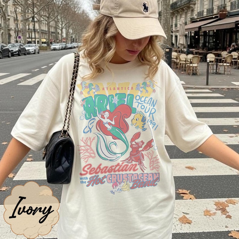May include: Ivory-colored t-shirt featuring a graphic of Ariel from The Little Mermaid, with the text "Ariel Ocean Tour 1991." The design includes illustrations of Ariel, Flounder, and Sebastian. The shirt has short sleeves.