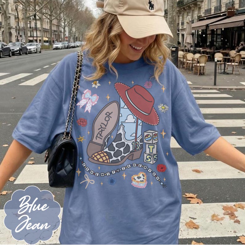 May include: A blue t-shirt featuring a graphic print of a cowboy boot with a cow print pattern, a red hat, the name Taylor, the number 13, and stacked blocks with the letters T, S, and S. The design includes small stars, bows, and a beaded string.