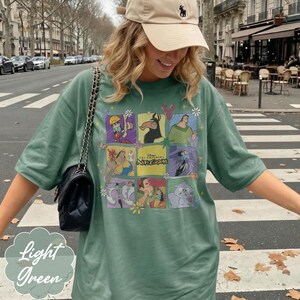 May include: A sage green t-shirt featuring a grid of colorful illustrations of characters from The Emperor's New Groove, including Kuzco, Pacha, and Yzma, with the movie title text in the center, paired with a beige baseball cap and a black bag.