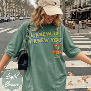 May include: A sage green t-shirt featuring the yellow text I KNEW IT, I KNEW YOU, a red heart with the letters TS, and the word STORY above the number 5 in yellow. The shirt is paired with a black quilted shoulder bag and a beige baseball cap.