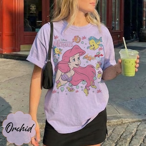 May include: A lilac t-shirt with a graphic of Ariel from Disney's The Little Mermaid. The design features Ariel with pink hair, a purple seashell bra, and various sea creatures. The shirt has short sleeves.