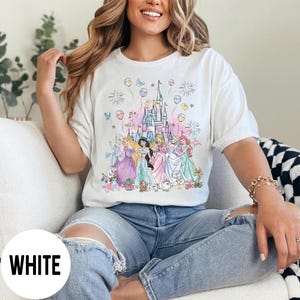 May include: White t-shirt featuring a colorful illustration of several princesses in front of a castle. The design includes balloons, fireworks, and floral accents. The word "WHITE" is printed at the bottom left.