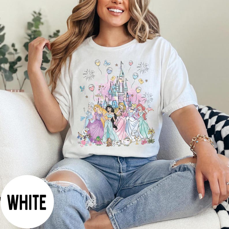 May include: White t-shirt featuring a colorful illustration of several princesses in front of a castle. The design includes balloons, fireworks, and floral accents. The word "WHITE" is printed at the bottom left.