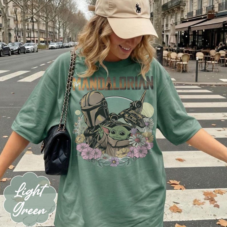 May include: Light green t-shirt featuring a graphic of The Mandalorian, Grogu, and a spaceship, surrounded by flowers. The word "MANDALORIAN" is printed across the top. The shirt is a light green color.