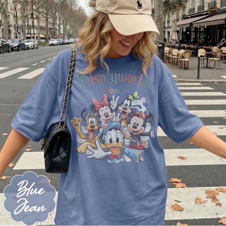 Pu&ograve; includere: T-shirt blu jeans con una grafica colorata dei personaggi Disney, tra cui Topolino, Minnie, Paperino e Pippo. Il testo "Disneyworld 2026" &egrave; stampato sopra i personaggi. La maglietta ha maniche corte.