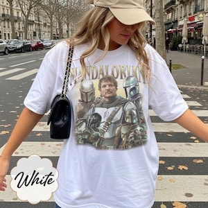 Pu&ograve; includere: Una t-shirt bianca a maniche corte con una stampa grafica di personaggi di The Mandalorian, tra cui una figura centrale e diverse figure in armatura. Il testo "MANDALORIAN" &egrave; stampato in un carattere color oro sopra l'illustrazione.